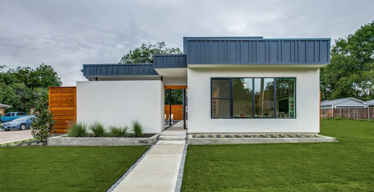 Modern flat-roof house exterior with stucco finish, black window frames, and wood fencing.