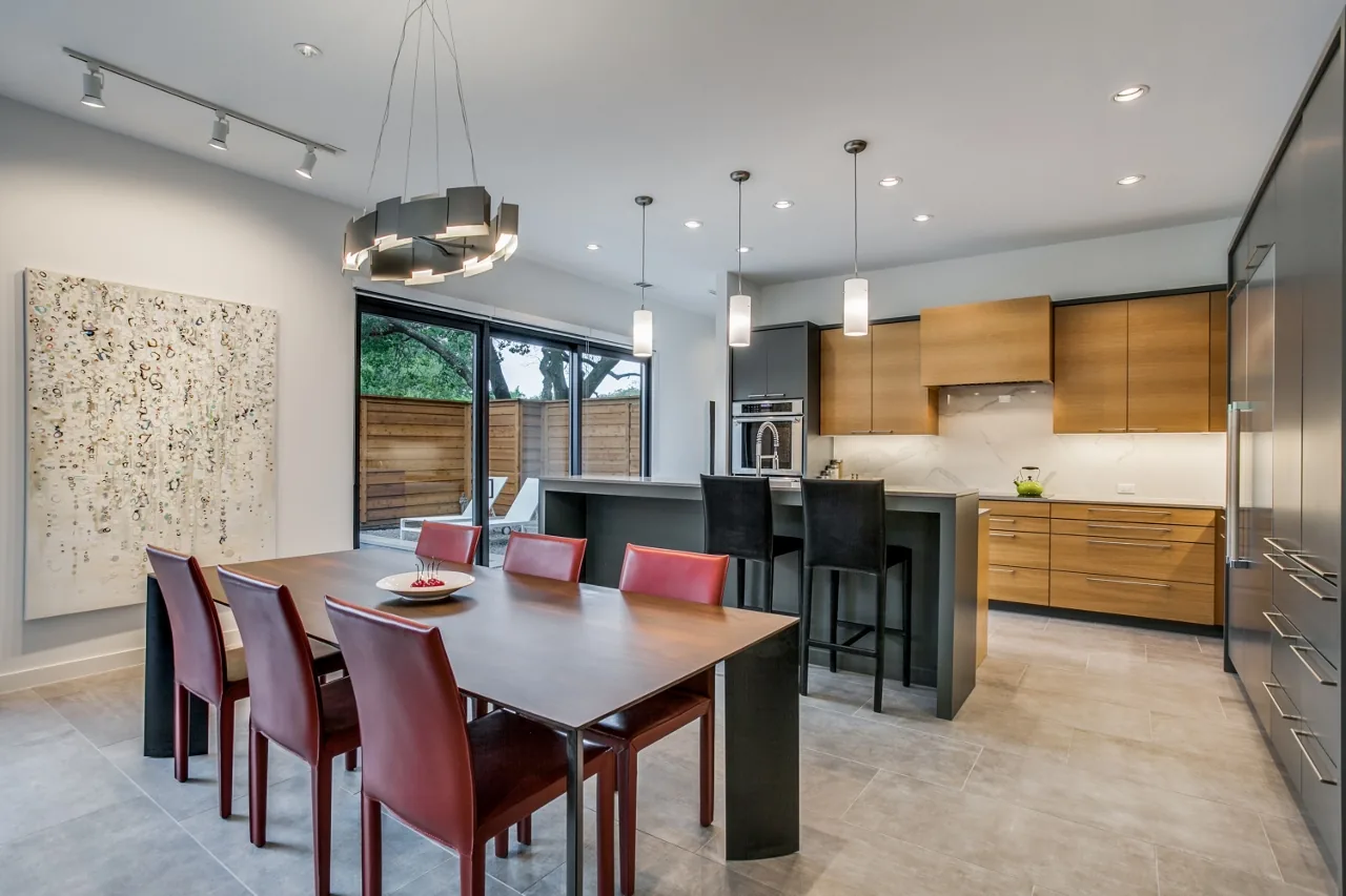 Modern dining room with a rectangular wood table and red chairs next to a kitchen featuring wood cabinets and island seating.