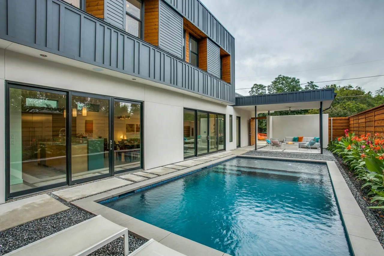 Modern backyard with a rectangular pool, glass sliding doors, and a covered patio featuring metal mesh chairs and turquoise pillows.