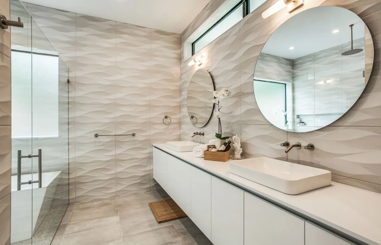 Bathroom with textured wall tiles, dual sinks under round mirrors, and an adjacent glass shower area with a frosted window.