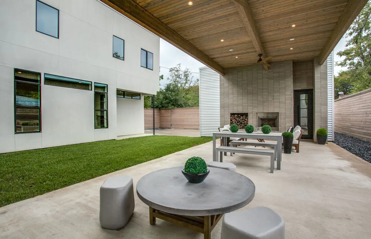 The Ribbon House outdoor living area features a wood beam ceiling-covered patio, a fireplace, and modern seating beside a grassy yard.