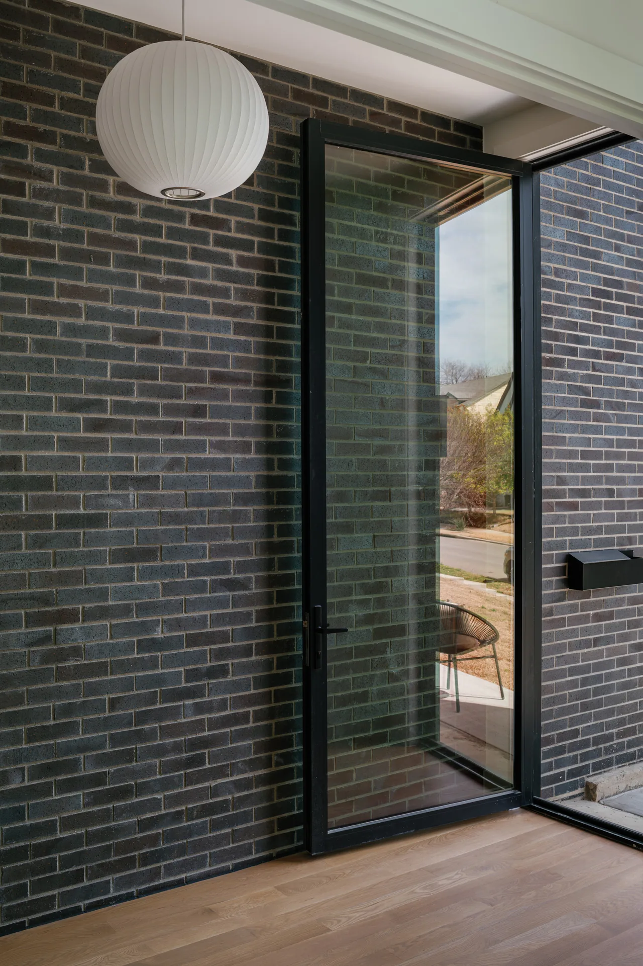 A black-framed sliding glass door opens to an outdoor area from an interior with dark brick wall and wood flooring.