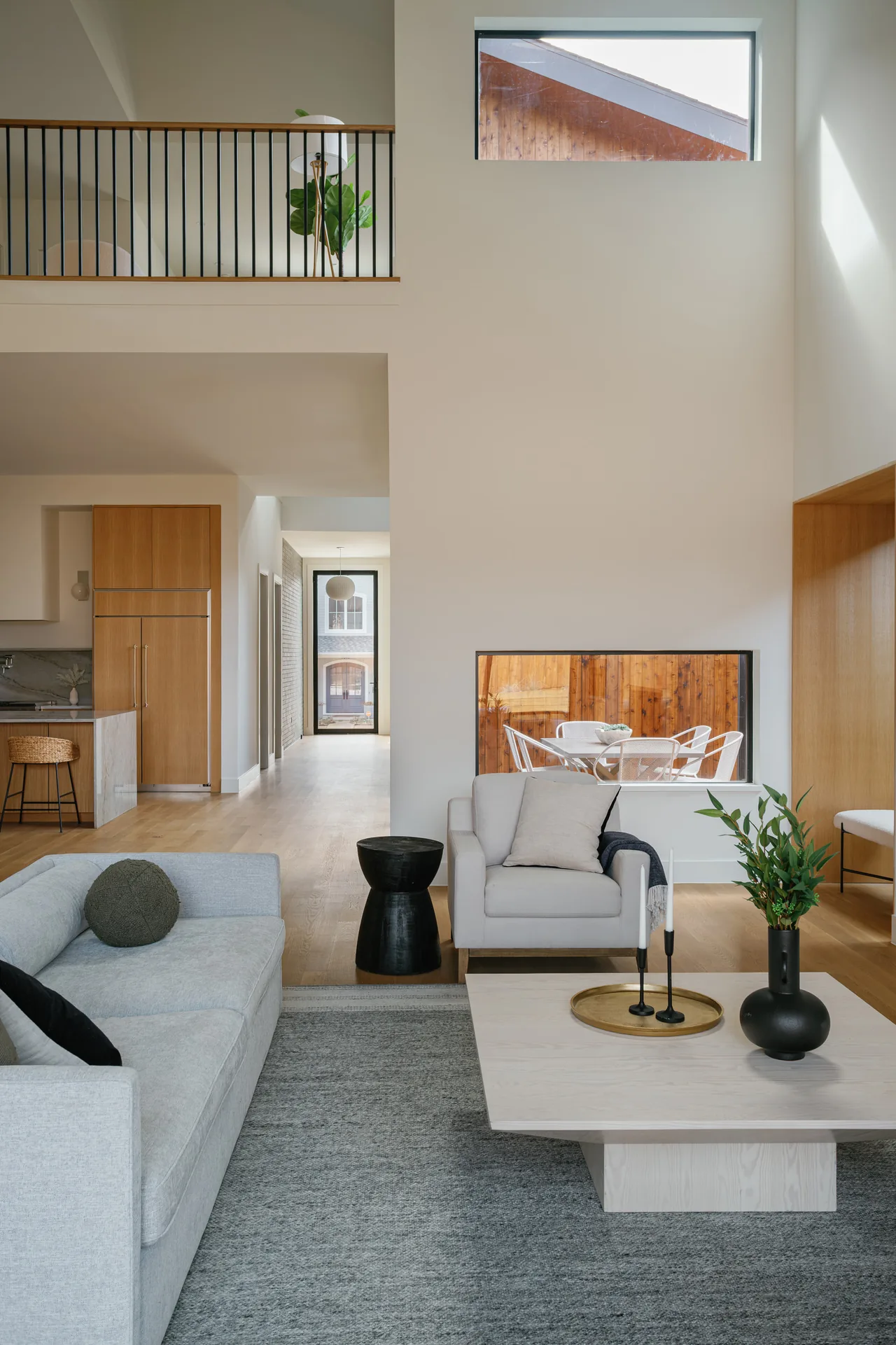 Living room with high ceilings and light wood floor opening into kitchen and hallway with views to outdoor dining.