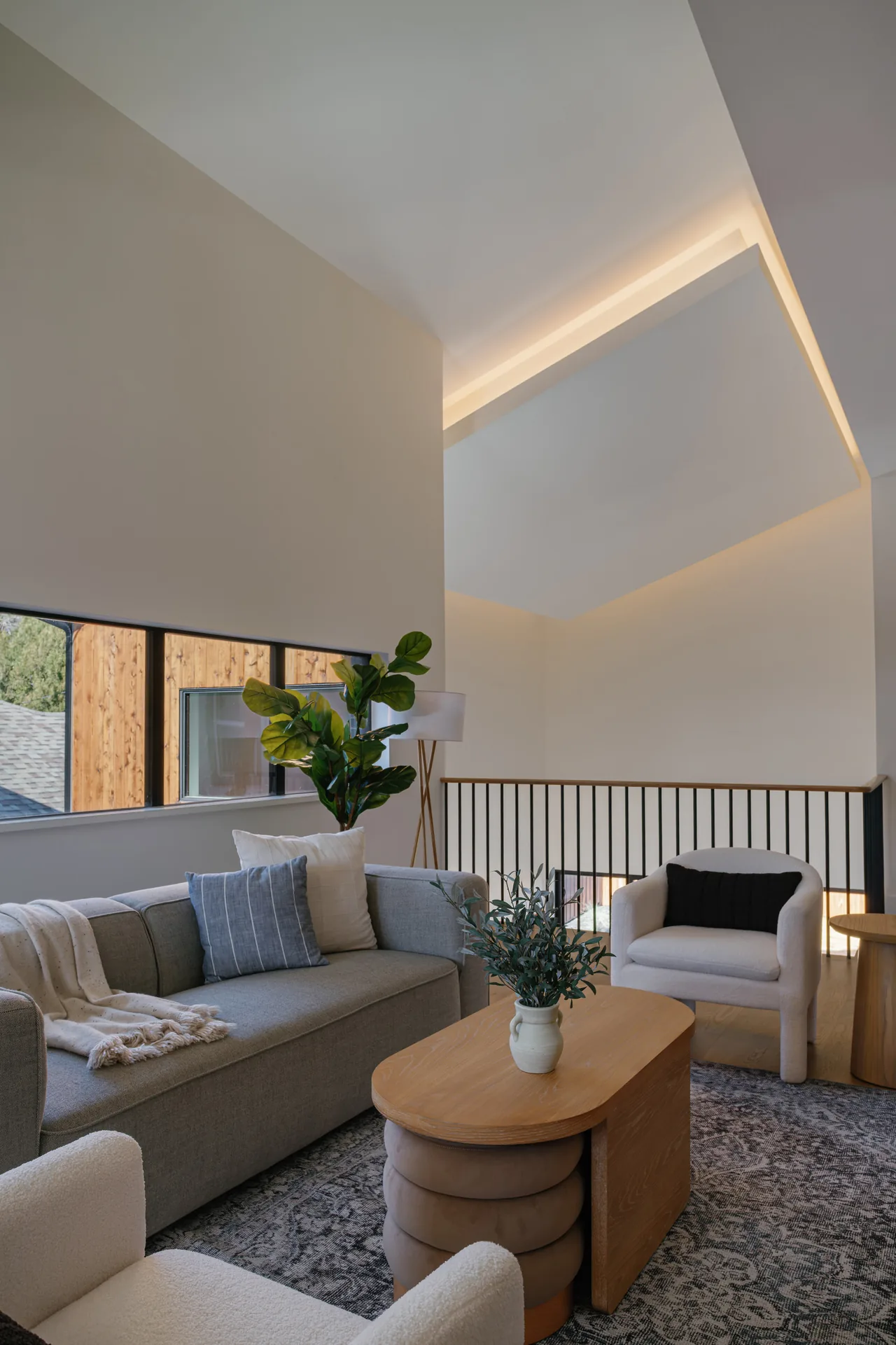 Living room with high ceilings and indirect lighting, furnished with a gray sofa, two white armchairs, and a wood coffee table with a plant.