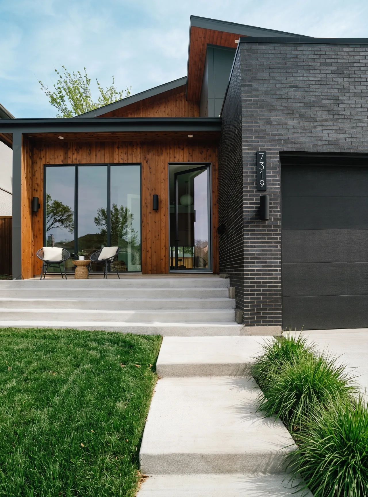Casa Loma by F+E Architects features a black brick garage with wood-paneled entry and concrete steps with outdoor seating.