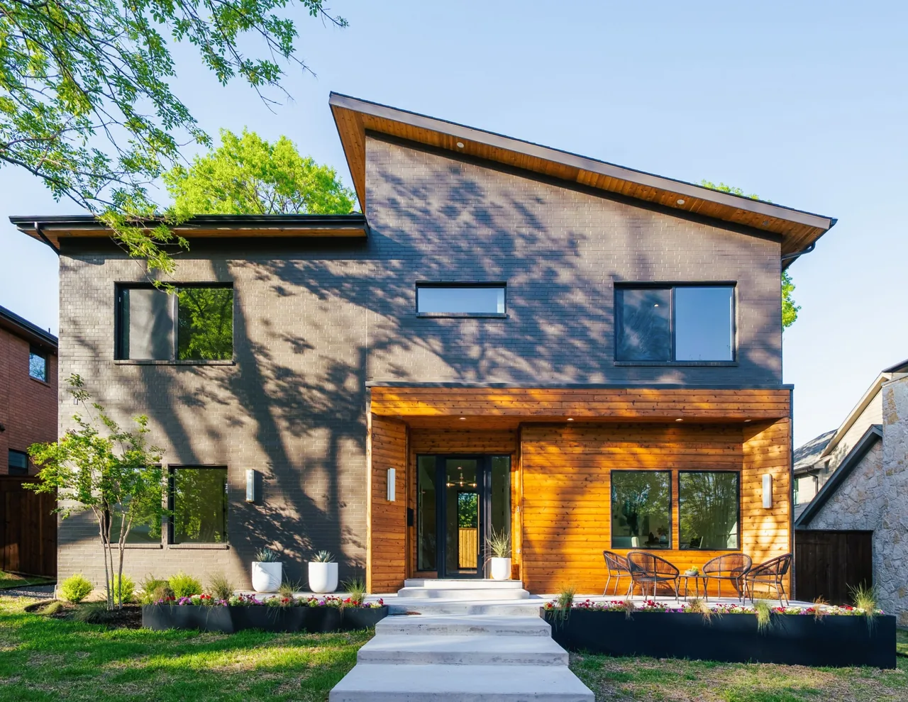 Front view of a modern two-story house with gray brick and wood siding, featuring a sloped roof and outdoor seating on the patio.