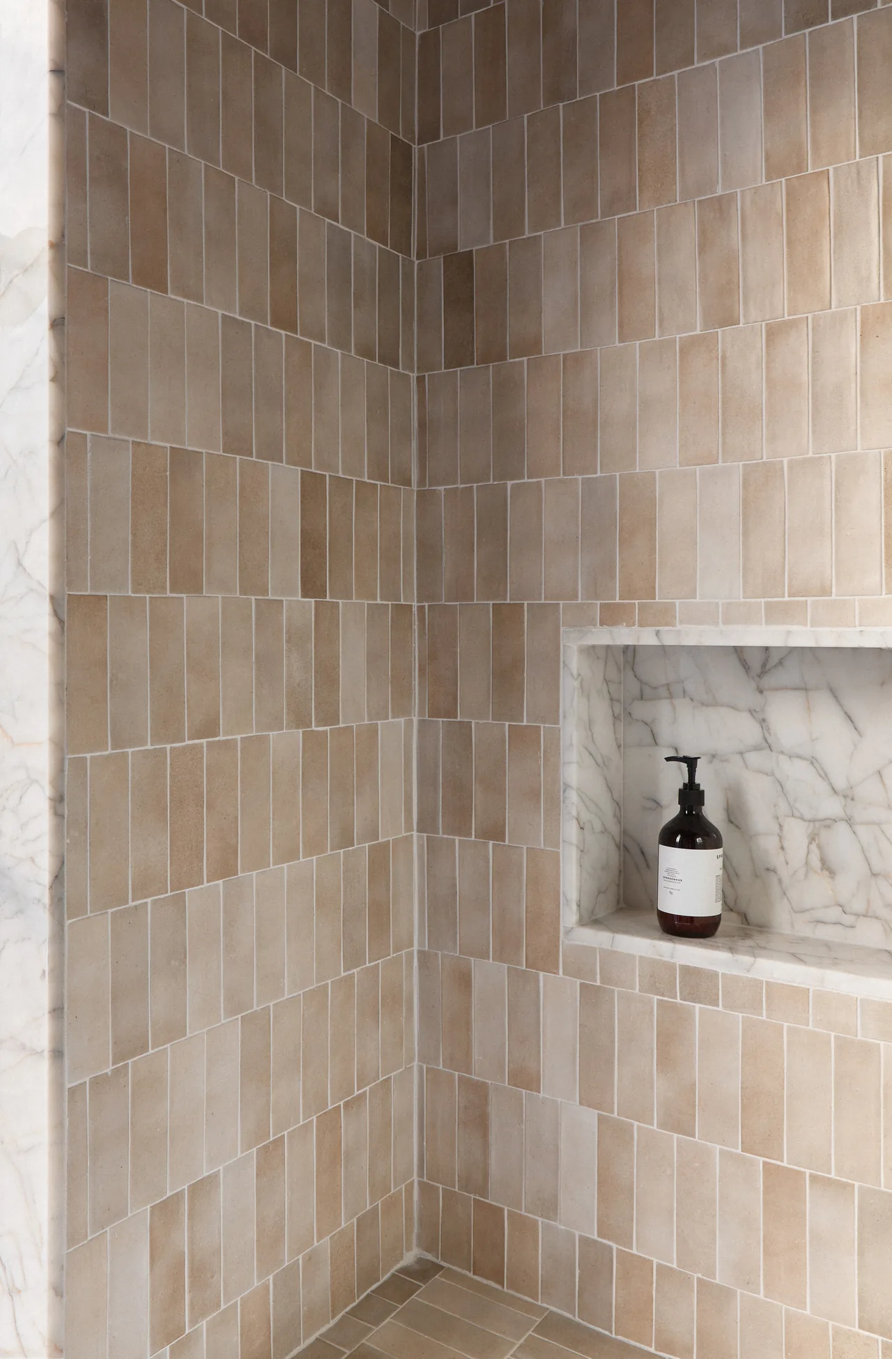 Shower corner features beige vertical rectangular tile walls and a marble niche with a soap dispenser.