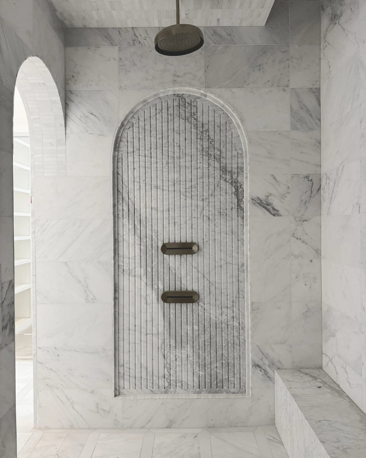 A marble shower features an arched niche with vertical grooves and a ceiling-mounted rain showerhead.