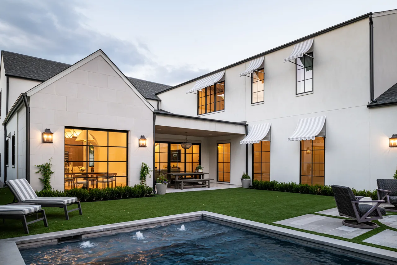 Modern two-story home exterior features black window frames, striped awnings, and a backyard pool with seating areas.