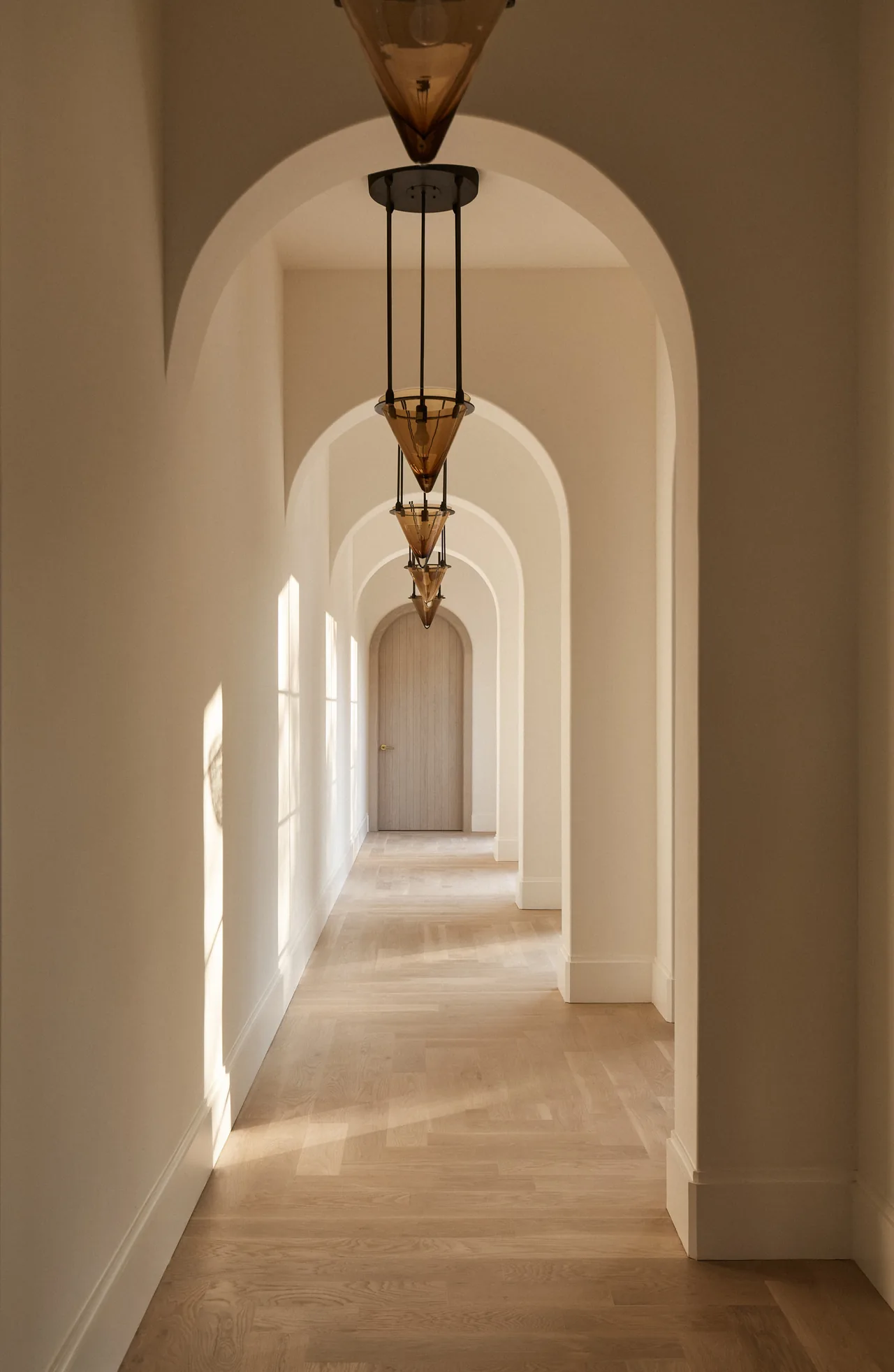 Hallway featuring arched doorways and patterned light wood flooring illuminated by conical pendant lights.