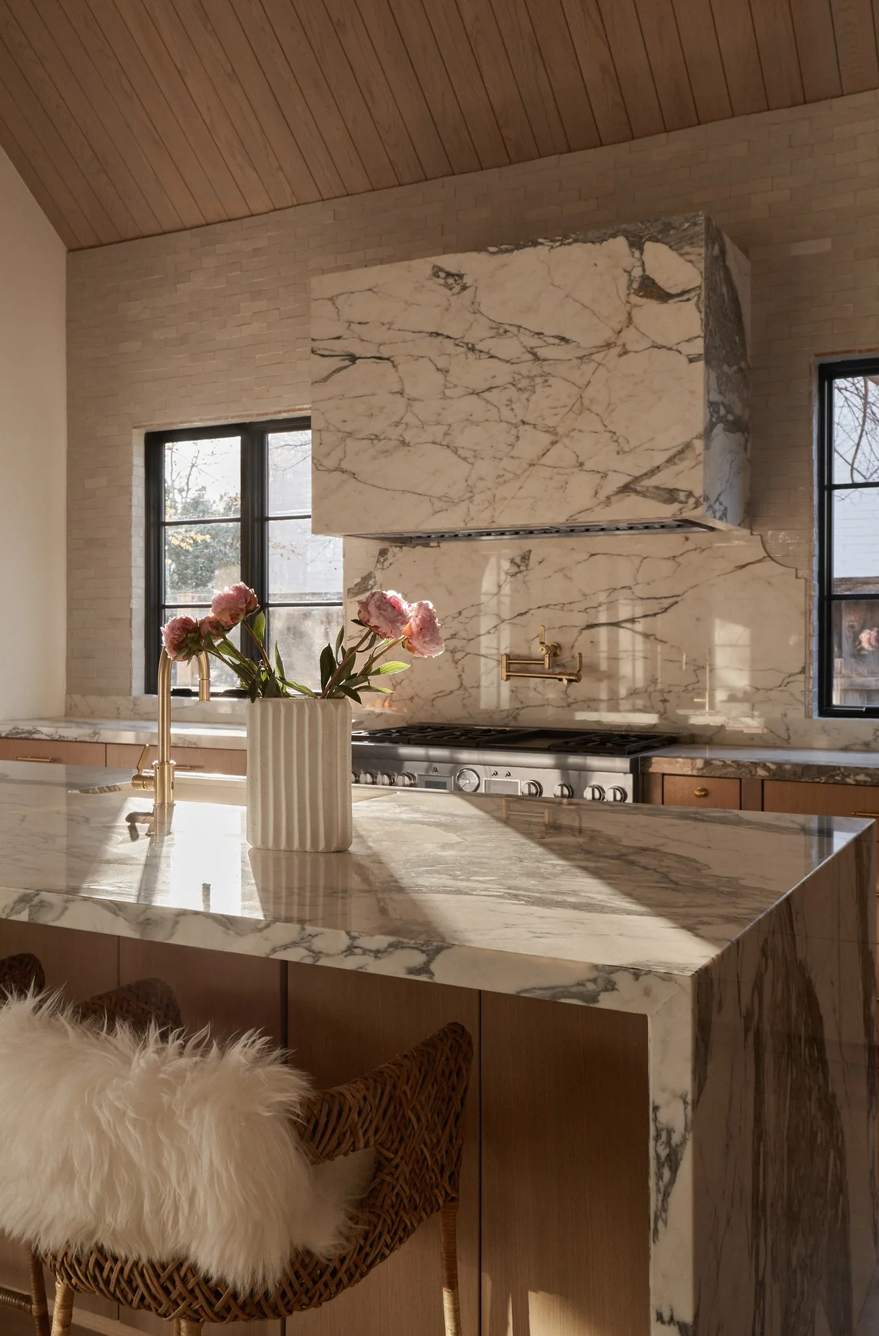 Kitchen featuring marble island and backsplash, wood cabinets, rattan stools with fur cushions, and brass fixtures.
