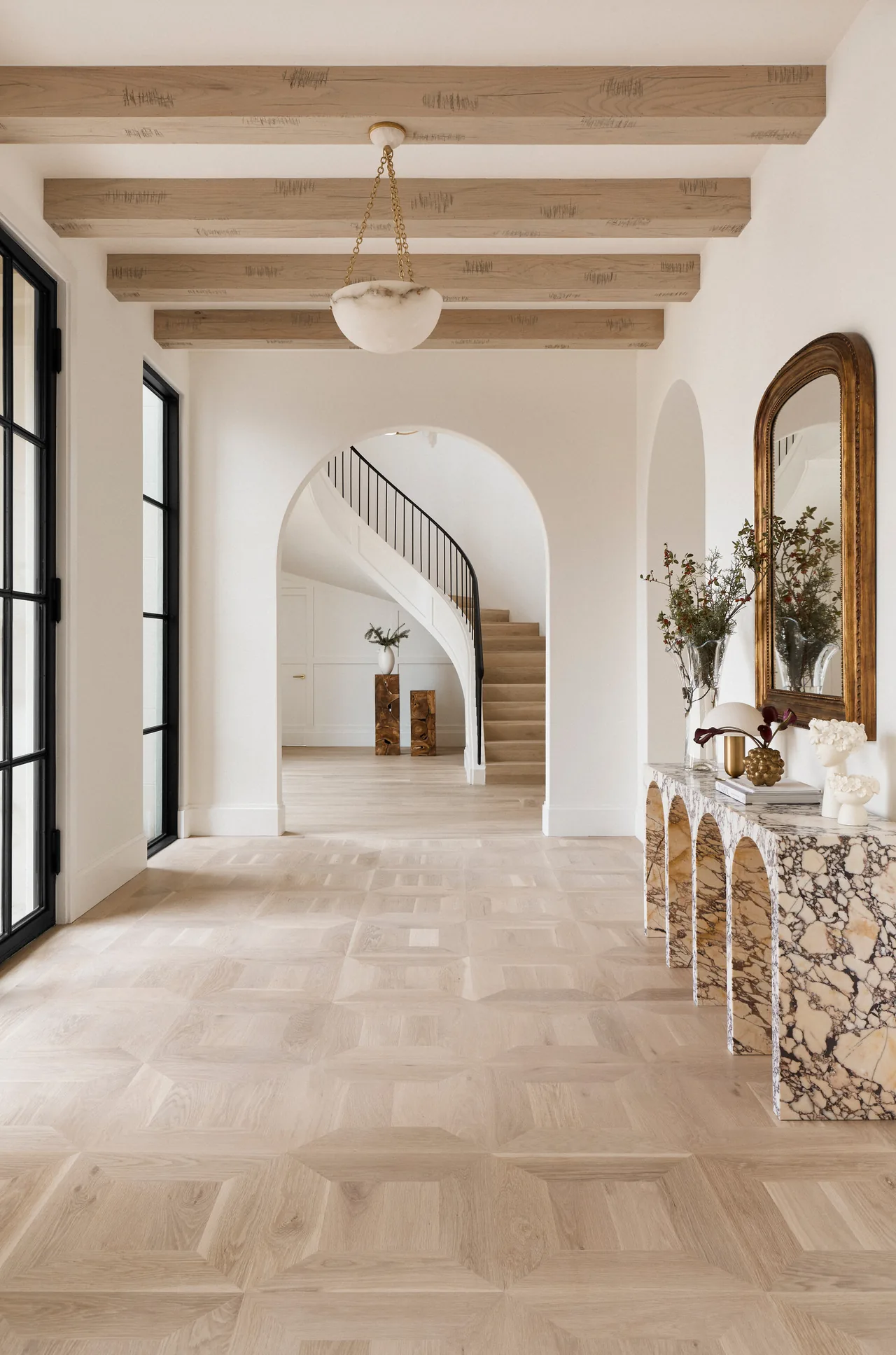 Entry hallway features an intricate light wood floor, exposed ceiling beams, arched openings, and a marble console table with decorative objects.