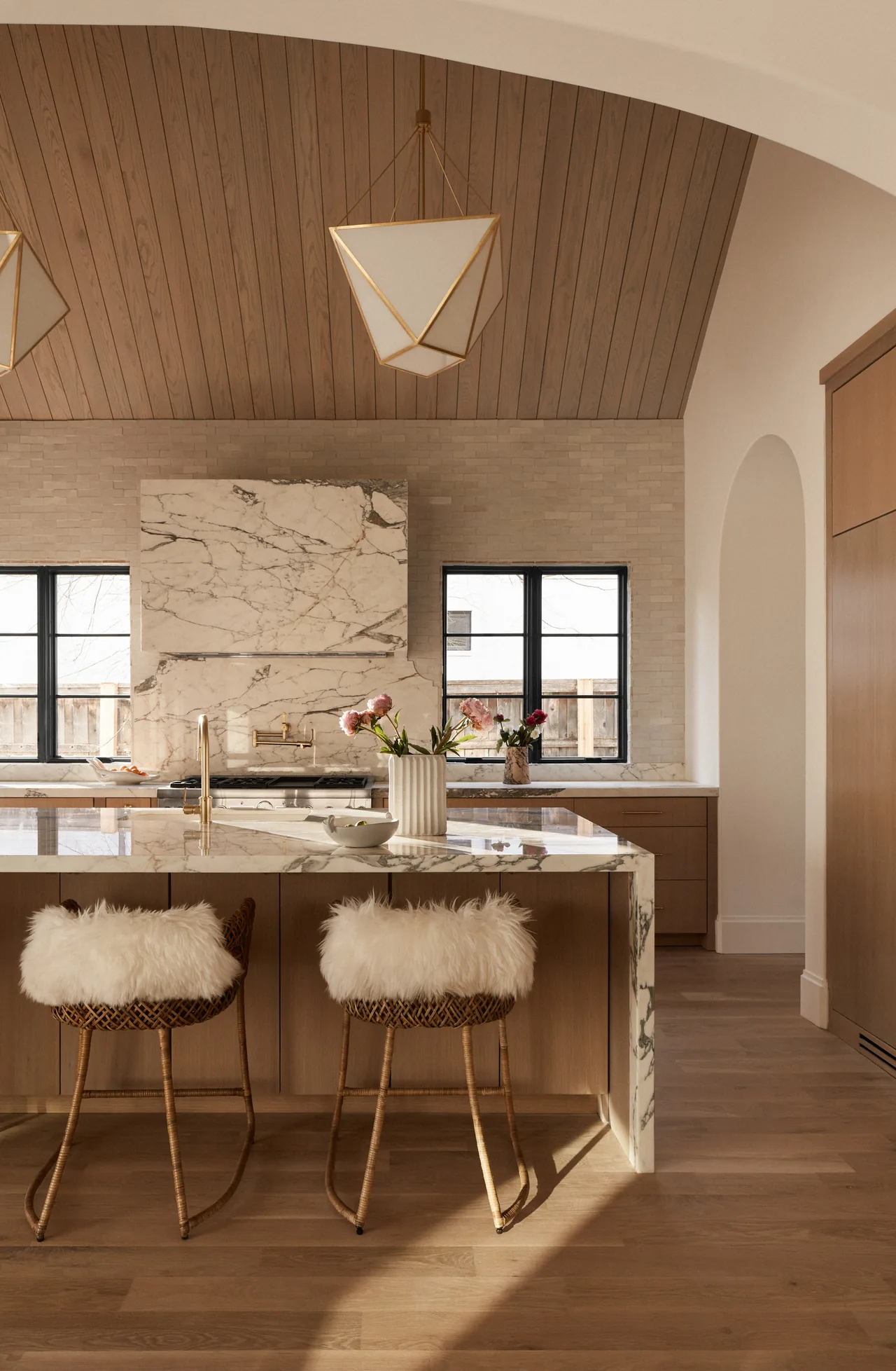 Kitchen features marble countertops and backsplash, vaulted wood ceiling, and two rattan stools with white fur cushions.