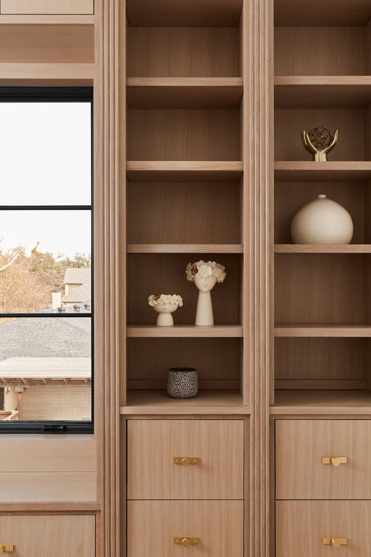 Custom built-in wooden shelving and drawers with gold hardware frame a black window grid, displaying decorative objects.