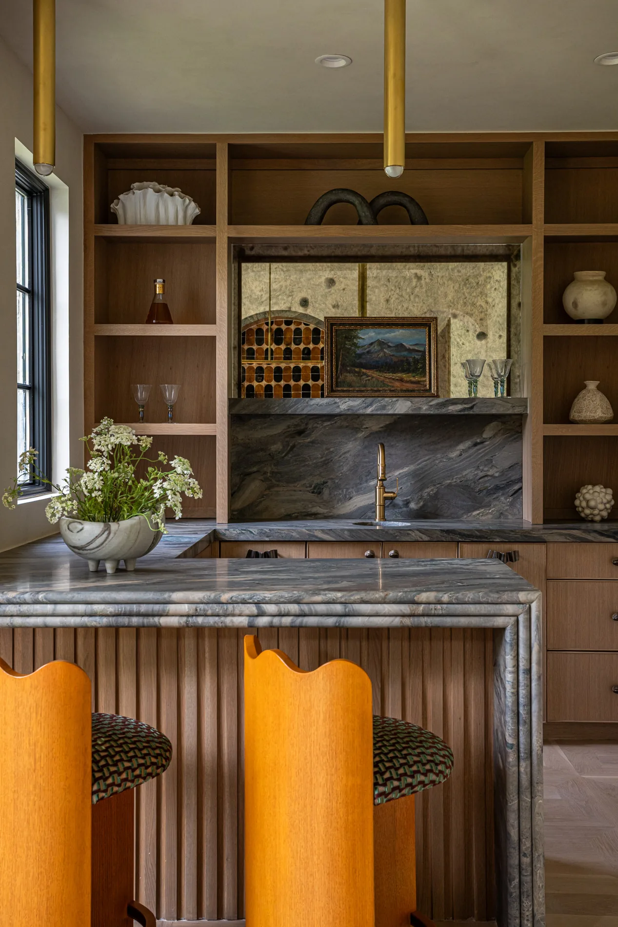 This kitchen features a gray marble island, wooden built-in shelves, and a brass faucet with decorative items and art.