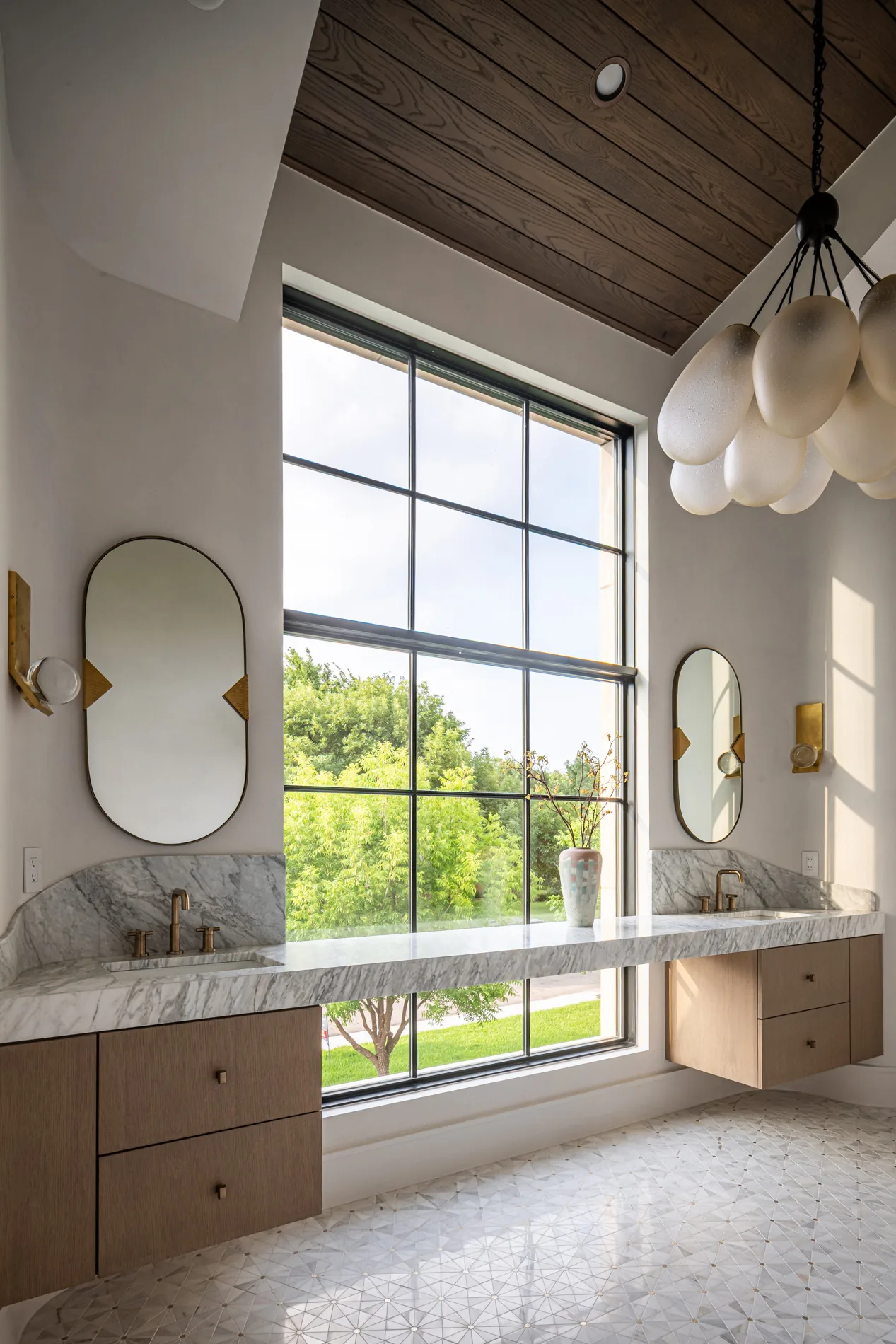 This bathroom includes a spacious double marble vanity with brass fixtures and a large window with grid framing.