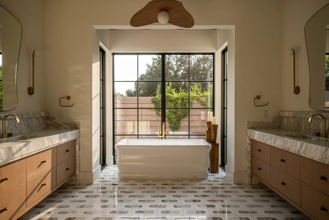 Spacious bathroom features a freestanding tub by large grid windows with brass fixtures and dual vanities with marble tops and wooden cabinets.