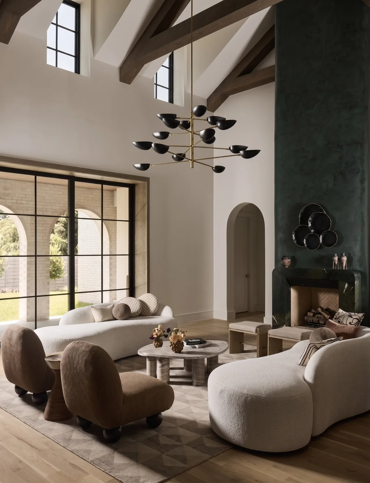 Living room interior featuring curved sofas, brown chairs, vaulted ceiling with exposed beams, and a large grid window overlooking an arched brick exterior.