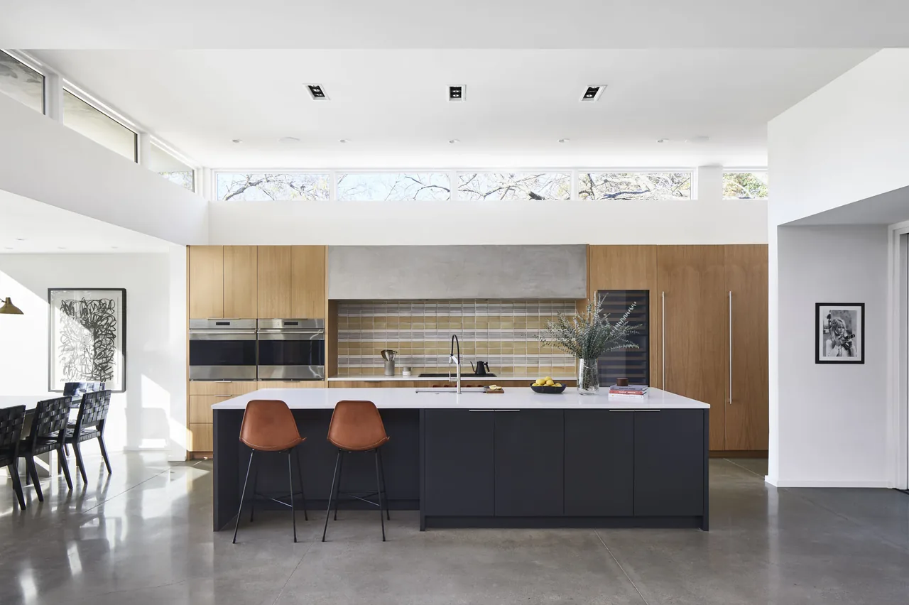 This modern kitchen features a dark island with white countertop, wood cabinets, clerestory windows, and built-in ovens.