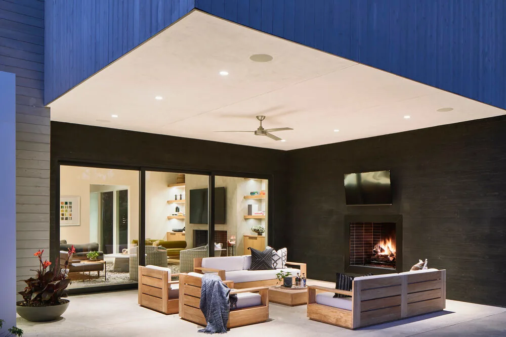 Outdoor living space features wooden seating, a fireplace, ceiling fan, and TV, connected to interior through sliding glass doors.