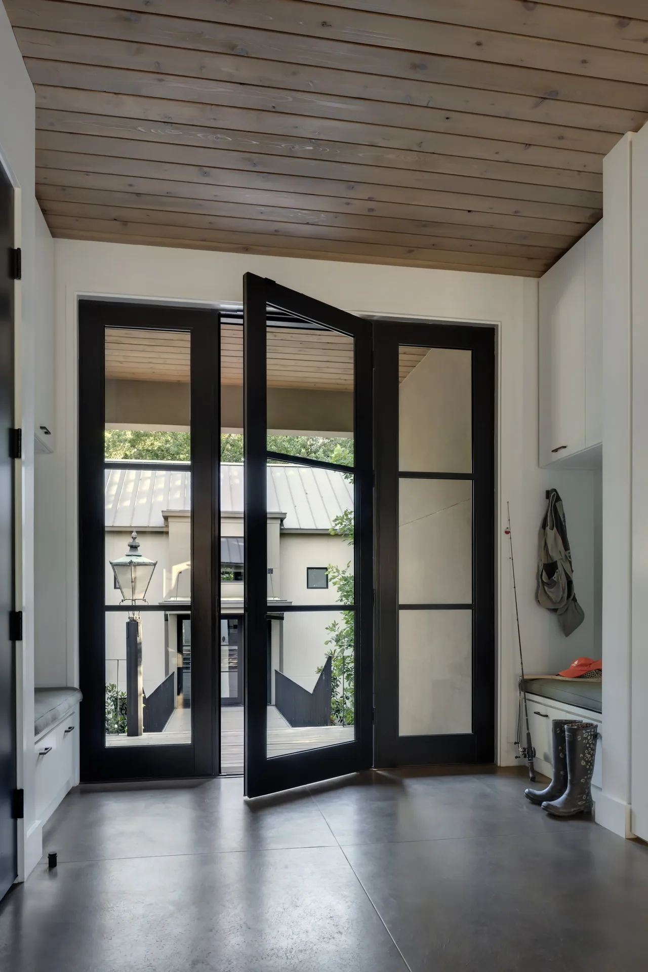 Mudroom area with black framed glass pivot door and bench seating, featuring gray concrete flooring and wood plank ceiling.