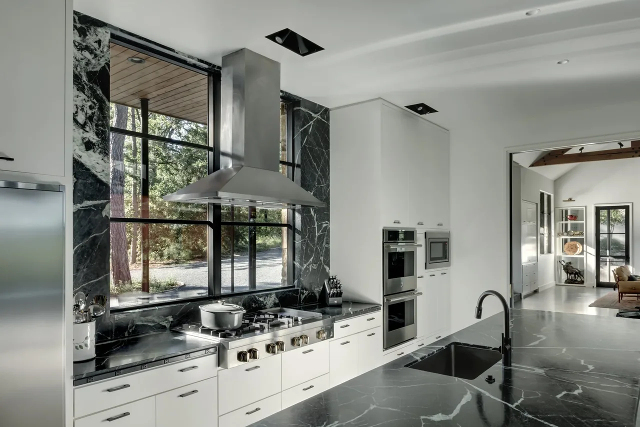 This kitchen at Pines Ranch features black marble countertops and backsplash, a gas range with stainless steel hood, and floor-to-ceiling black window frames.