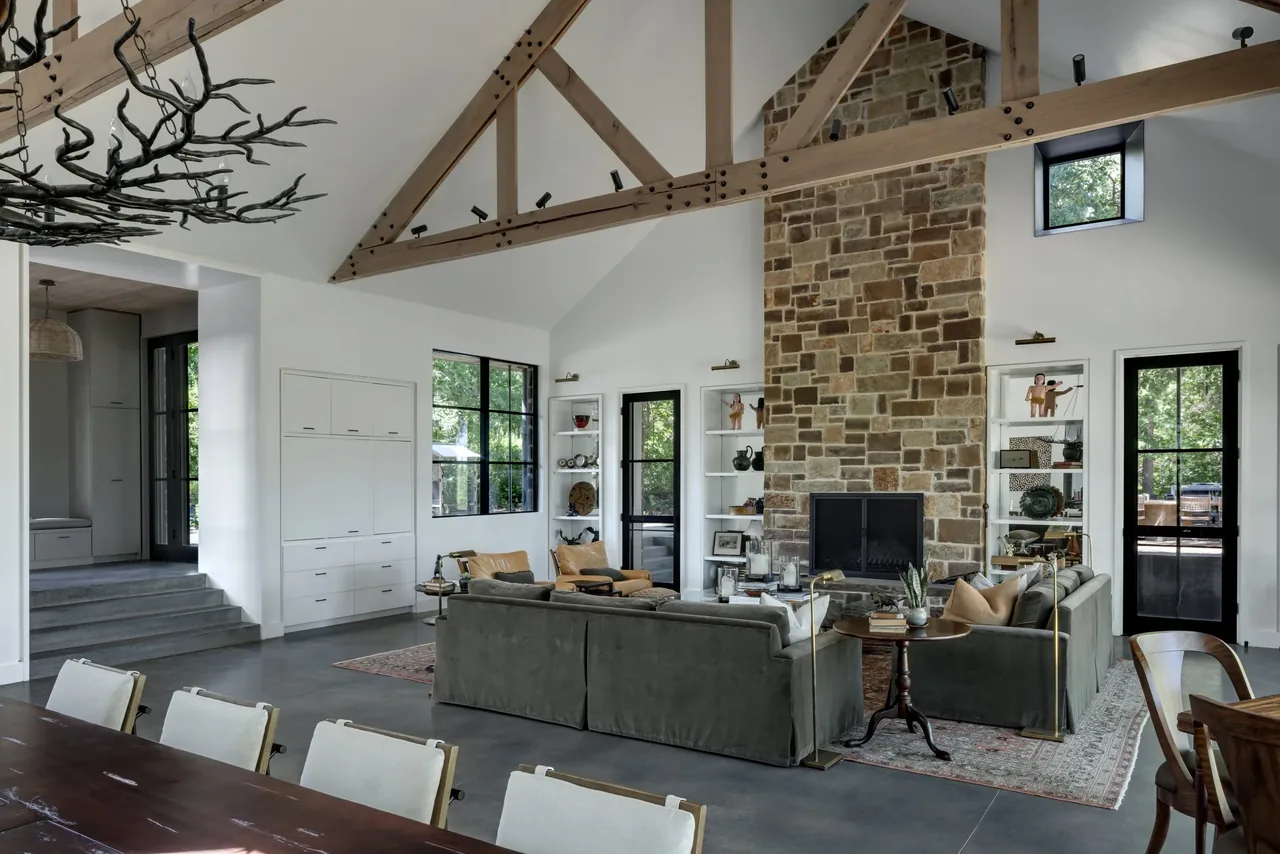 Living room with vaulted ceiling, exposed wood beams, stone fireplace, built-in shelving, and large windows with black frames.