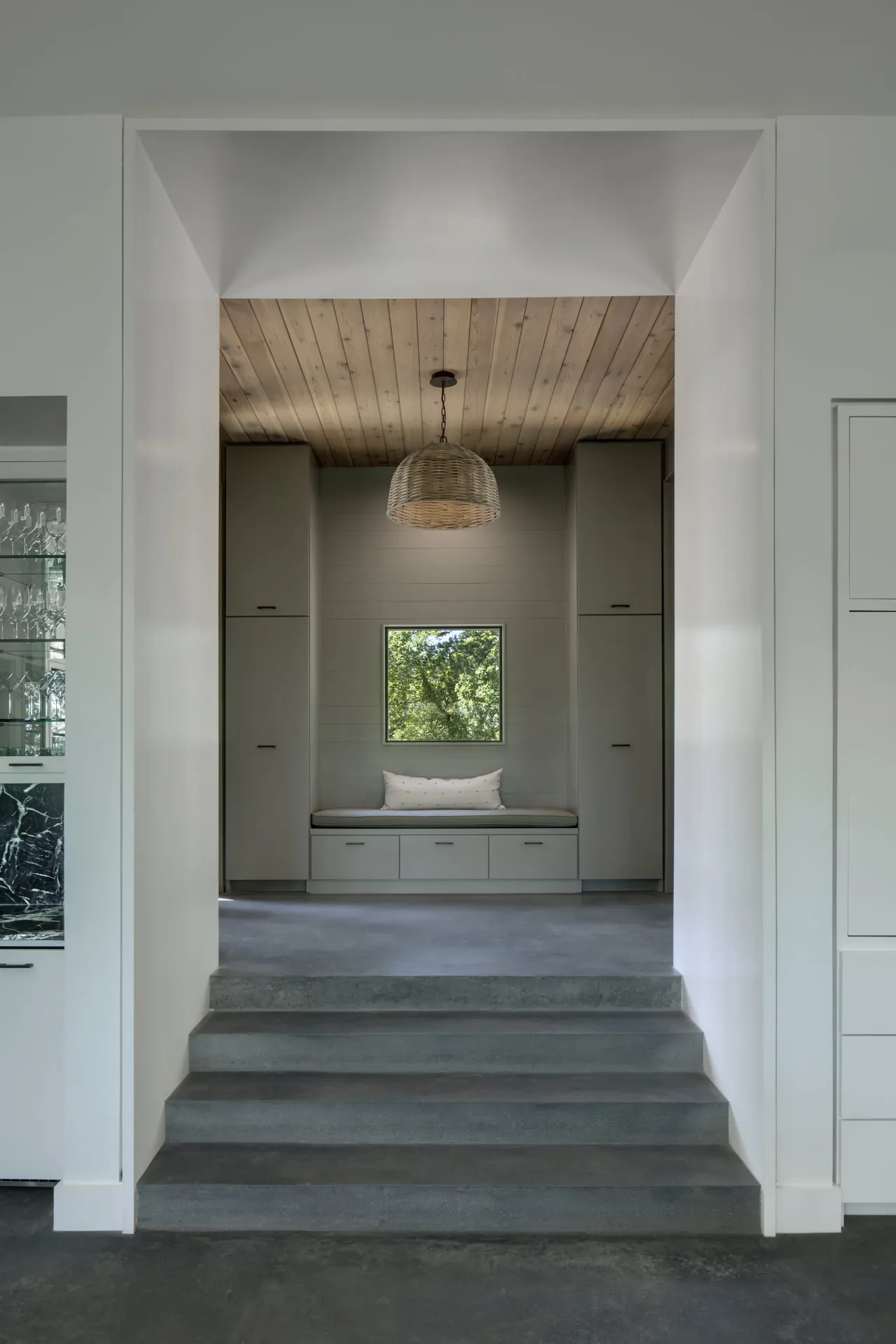 Built-in bench seating alcove below a wood plank ceiling and a woven pendant light, accessed by concrete steps
