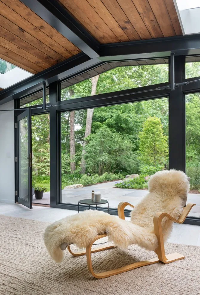 Living area with a lounge chair covered in sheepskin and large black window frames opening to a green garden.