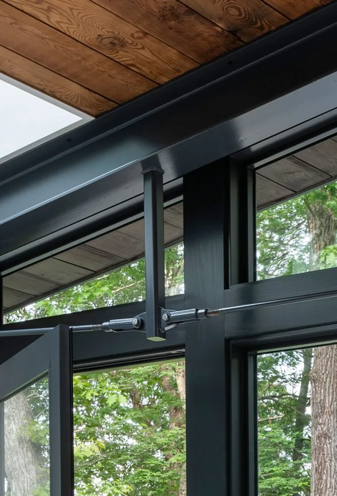Black steel-framed glass door and window with wood plank ceiling above, showing a view of trees outside.