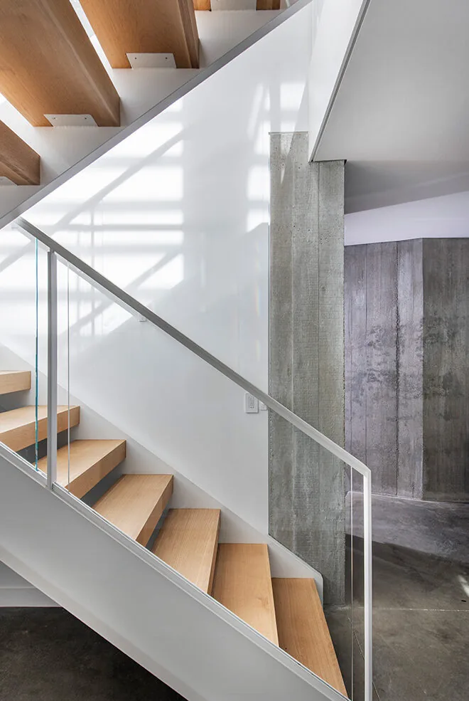 Floating wood stairs with glass railing are installed beside a textured concrete wall in a modern home.