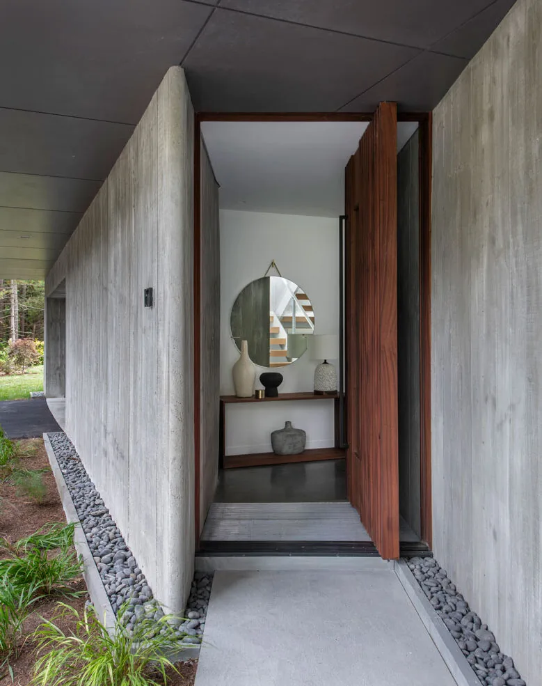 Modern home entry features a wooden pivot door within board-formed concrete walls and river rock edging.