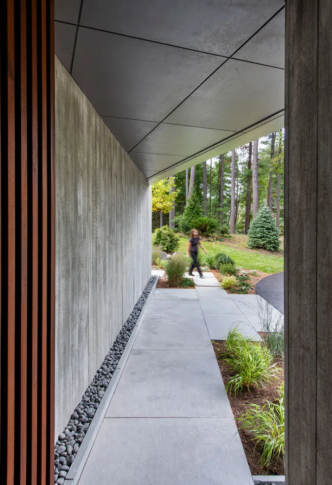 Covered concrete path alongside a textured concrete wall opens to a landscaped garden with trees.