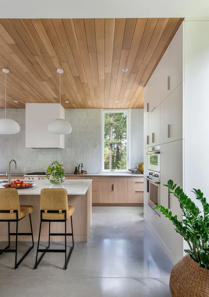 Kitchen features wood-paneled ceiling, concrete floor, and light wood cabinetry with a window revealing trees outside.