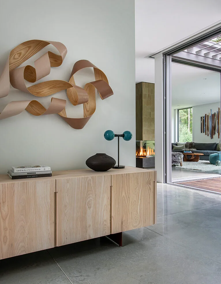 Modern living room with light wood sideboard and abstract wood wall sculpture, showing a glimpse of a fireplace and lounge area.
