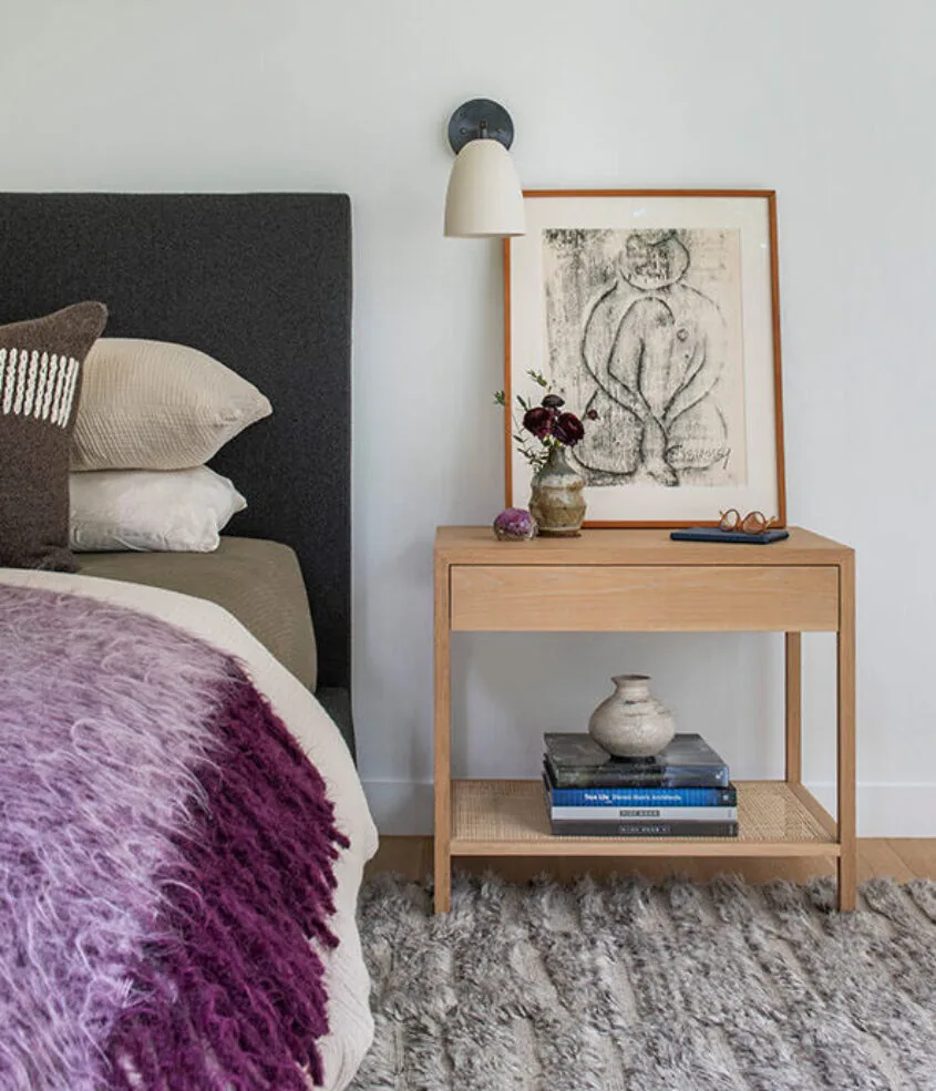 Bedroom corner featuring a wooden nightstand with decor, a textured rug, and a wall sconce above framed art.