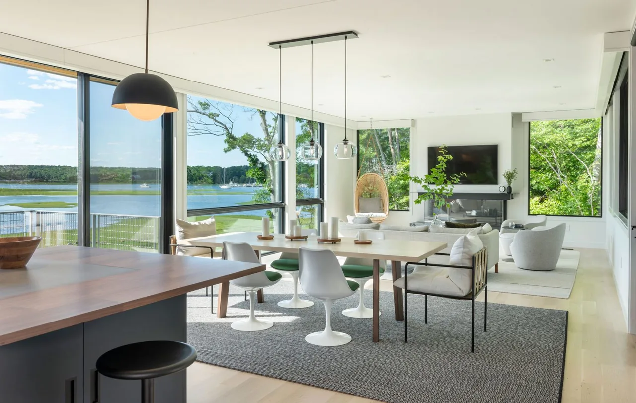 The open dining and living space includes a large kitchen island, modern seating, and expansive windows overlooking a tidal marsh.