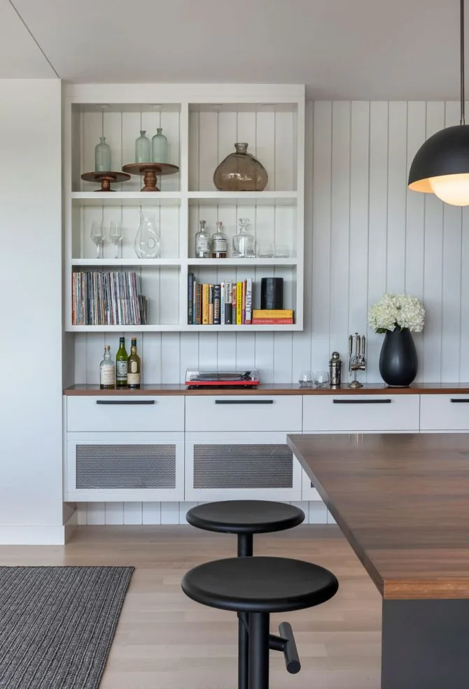 This kitchen has a wooden island with black stools and white cabinetry including open shelving displaying glassware and books.