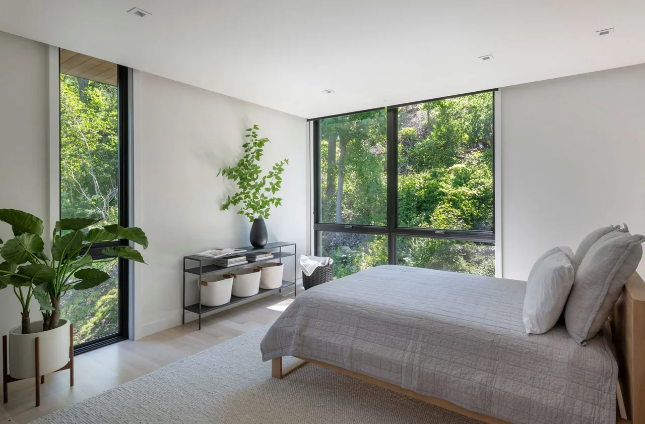 Bedroom with floor-to-ceiling black-framed windows offering a lush green view and minimalist décor.