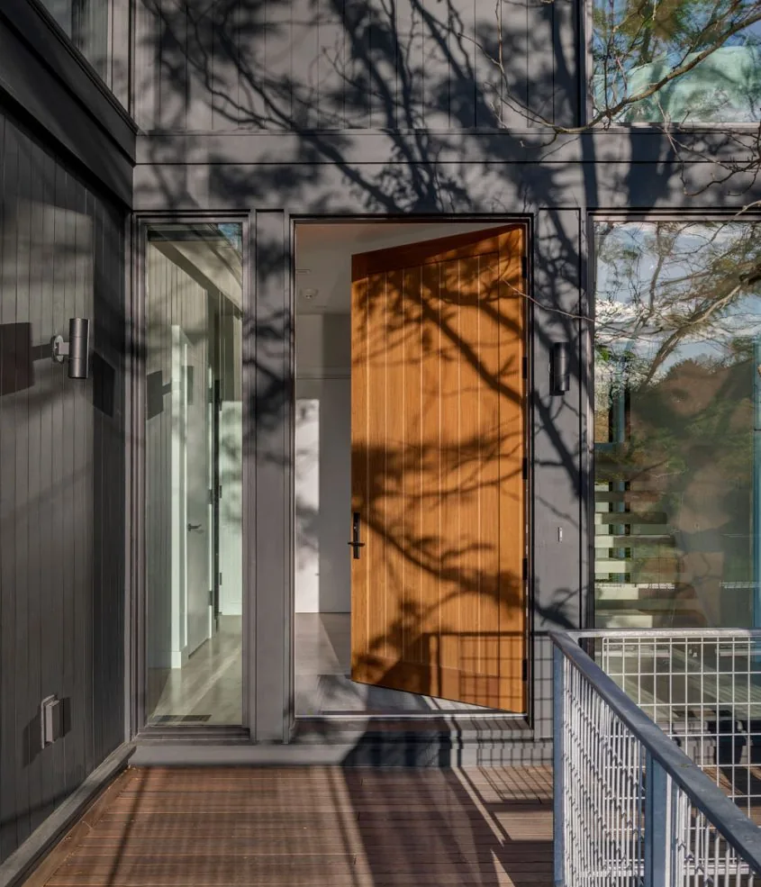 Exterior view of a modern home's entrance with a wooden pivot door and dark vertical wood siding at Tidal Marsh Overlook.