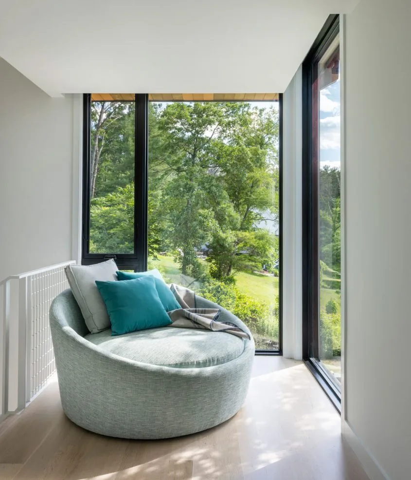 A curved lounge chair with blue and gray pillows by floor-to-ceiling windows offers a scenic view of trees and marsh outside.