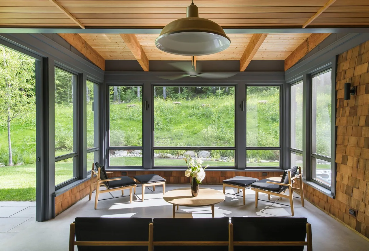 A porch seating area features wood-framed black chairs and a round table under a wooden beam ceiling with large windows overlooking greenery.