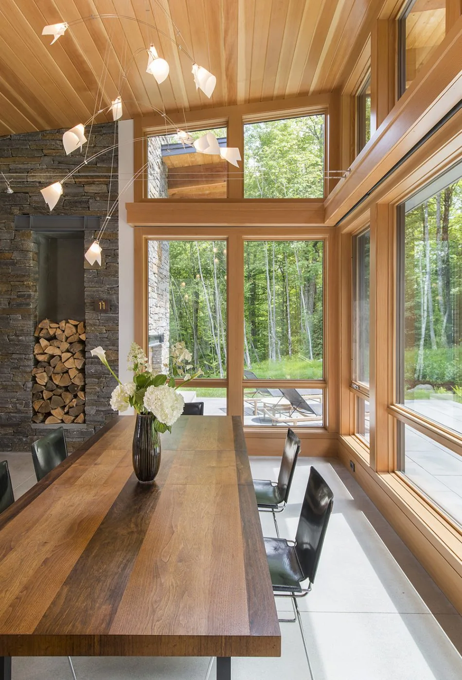 Dining room with a wood beam ceiling, stone wall storage for firewood, and expansive floor-to-ceiling glazing.
