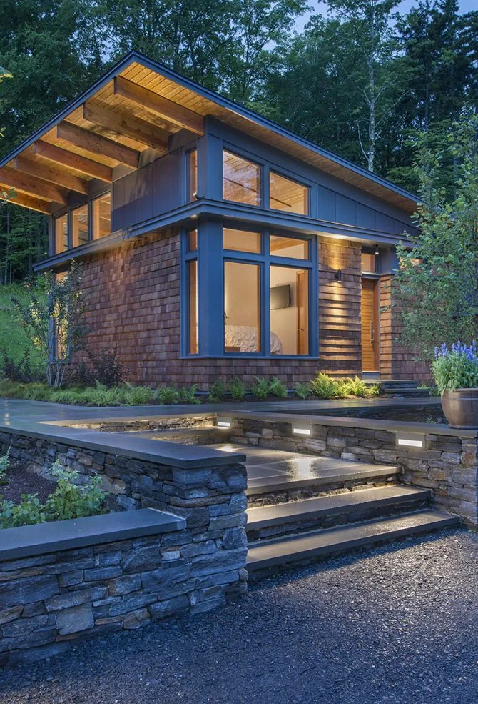 Modern residence with wood shingle siding and expansive windows illuminated at dusk, showing clearly defined landscaping and stone stairs.