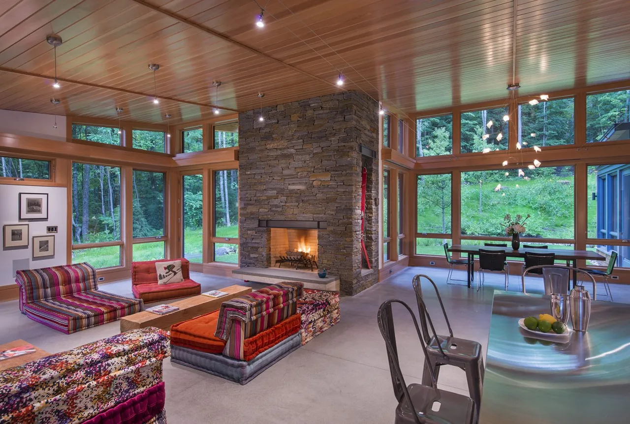 Modern living space with a stone chimney fireplace and large wood-framed windows overlooking greenery.