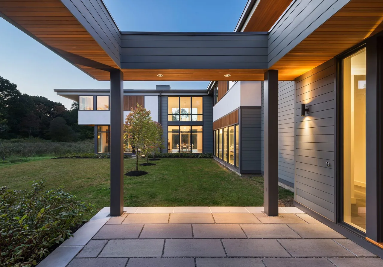 Covered patio facing the rear exterior of the Modern Blueberry Farm house with large windows and wood siding.