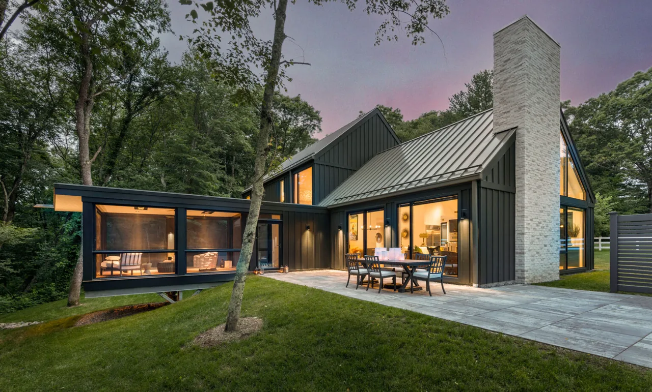 The Silver Hill House features a black wood siding exterior with a standing seam metal roof and a stone chimney, adjacent to a paved patio and lawn.