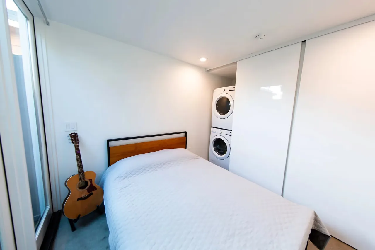 Bedroom featuring a bed with white bedding, an acoustic guitar, and a closet concealing a stacked washer and dryer.