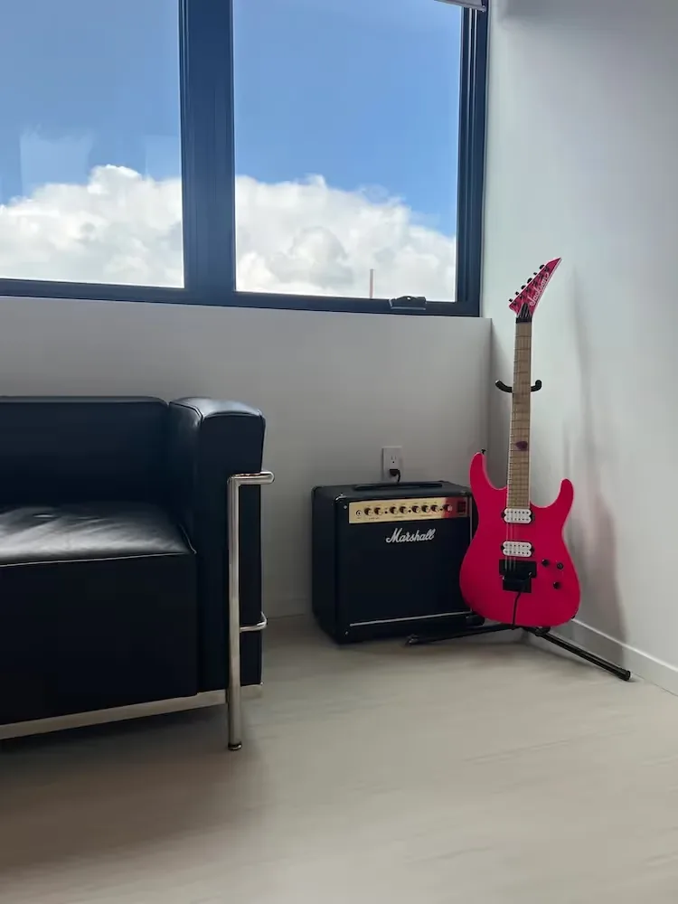 A black leather sofa sits beside a pink electric guitar and black amplifier under a window with roller blinds in a modern room corner.