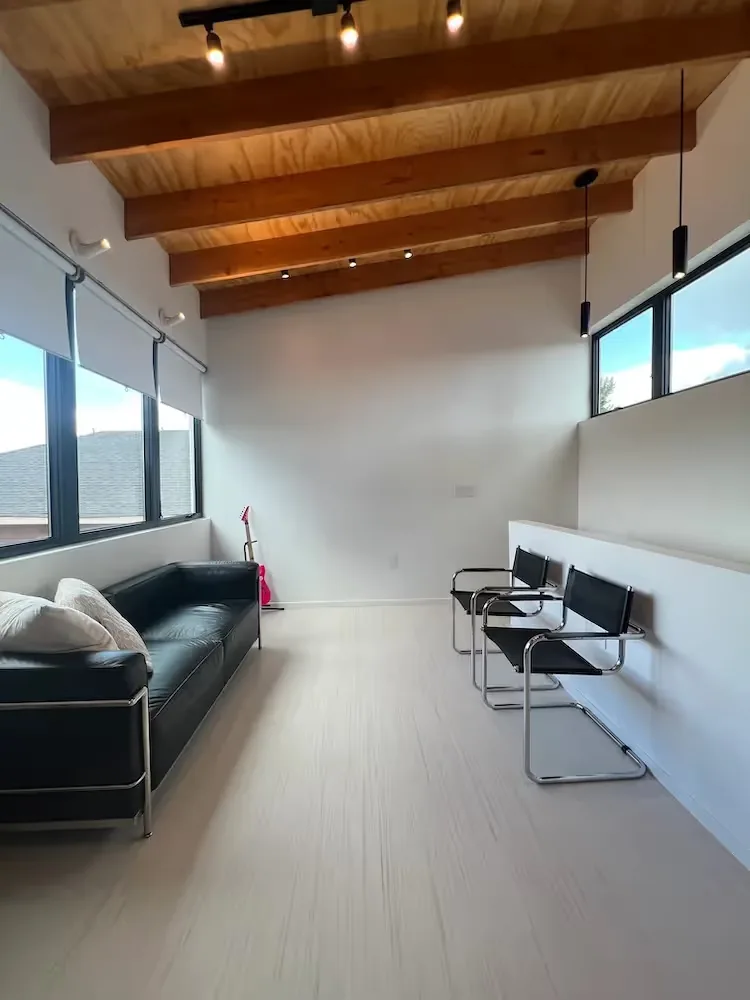 A minimalist living room features exposed wood beams, black leather furniture, and large windows with roller blinds.